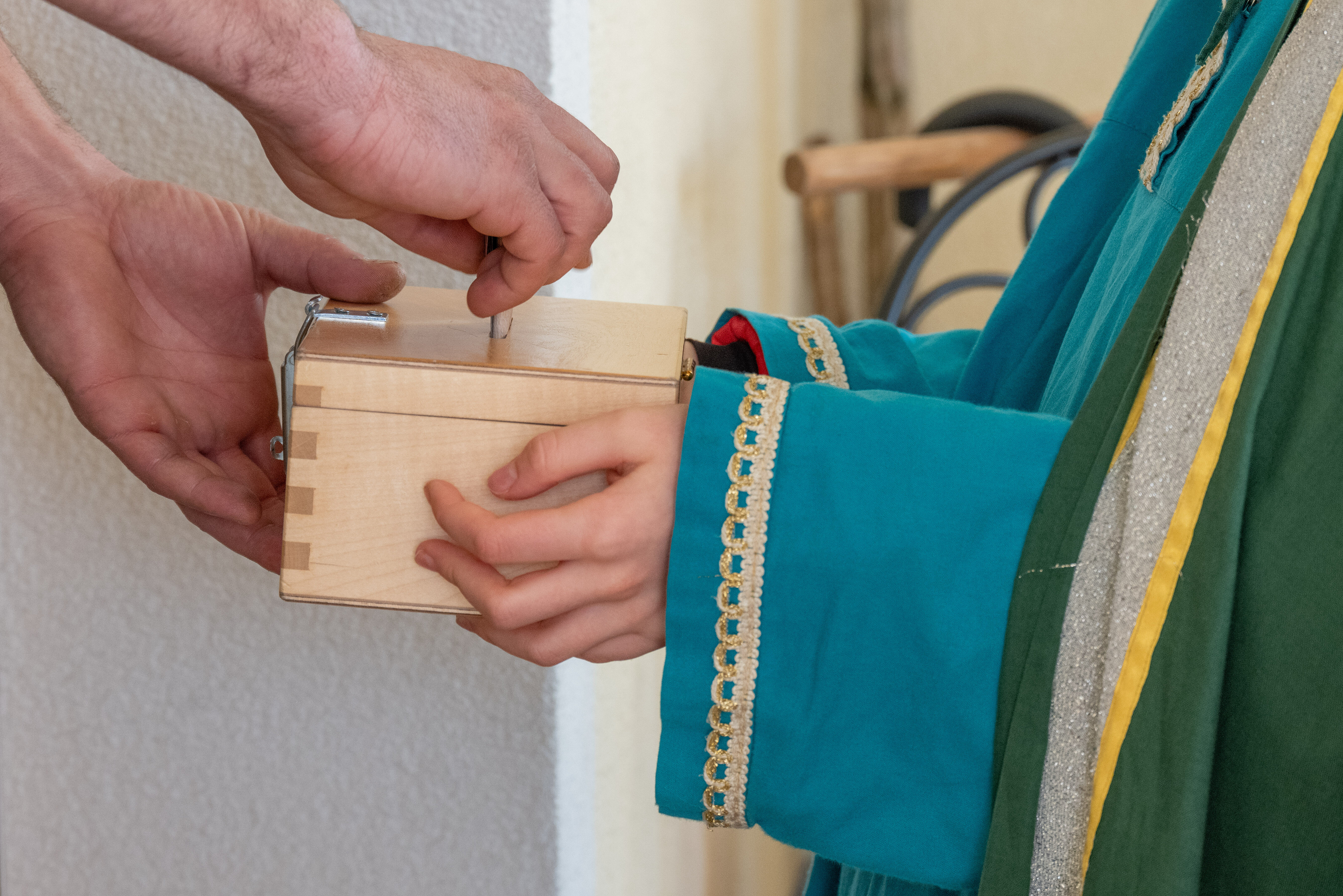 Das Bild zeigt eine Szene, in der eine Person Geld in eine kleine Holzkiste legt, die von einem Kind gehalten wird.