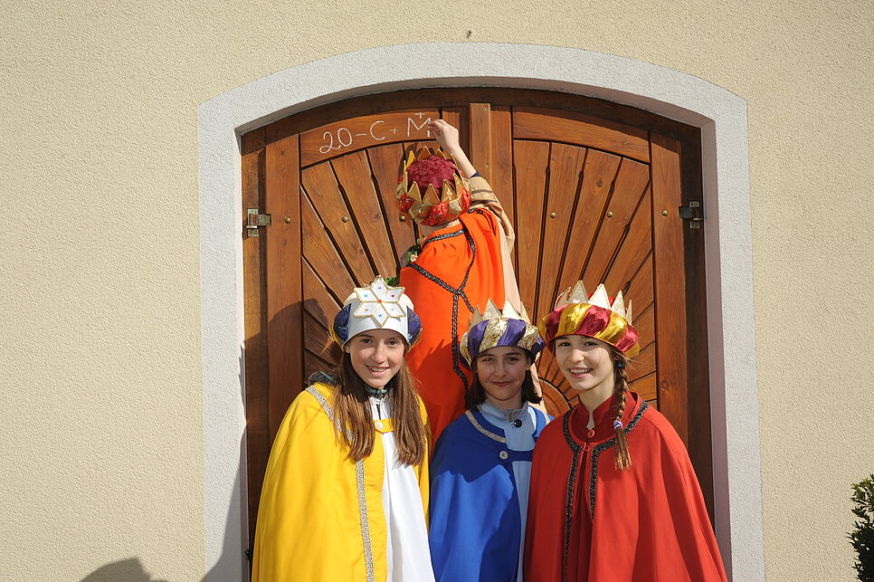 Vier Kinder, gekleidet als die heiligen drei Könige samt Sternsinger, schreiben an eine Tür.