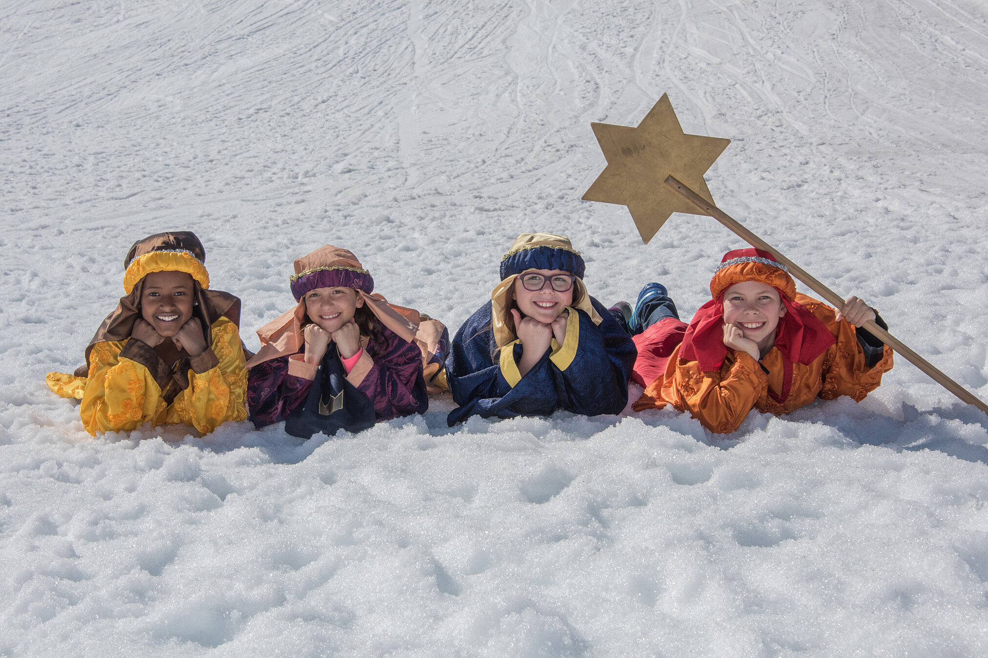 Die heiligen drei Könige samt Sternsinger liegen auf einer schneebedeckten Wiese und lachen in die Kamera.