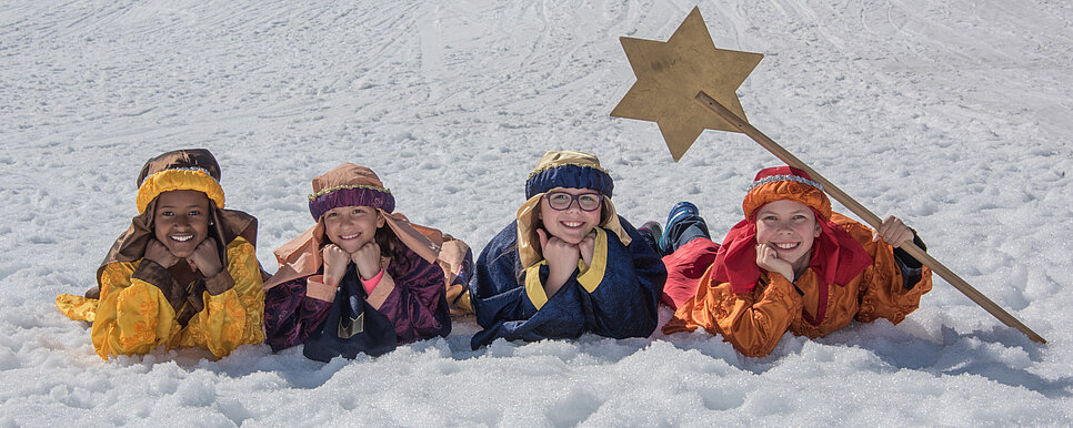Die heiligen drei Könige samt Sternsinger liegen auf einer schneebedeckten Wiese und lachen in die Kamera.