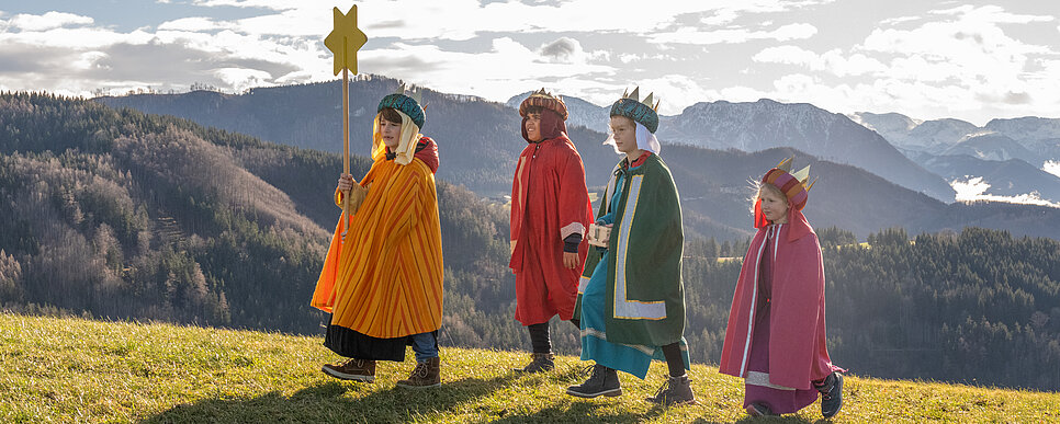 Vier Kinder in bunten Königsgewändern und Kronen gehen in einer Reihe über einen Hügel, das vorderste trägt einen großen Stern. Im Hintergrund erstreckt sich eine winterliche Berglandschaft.