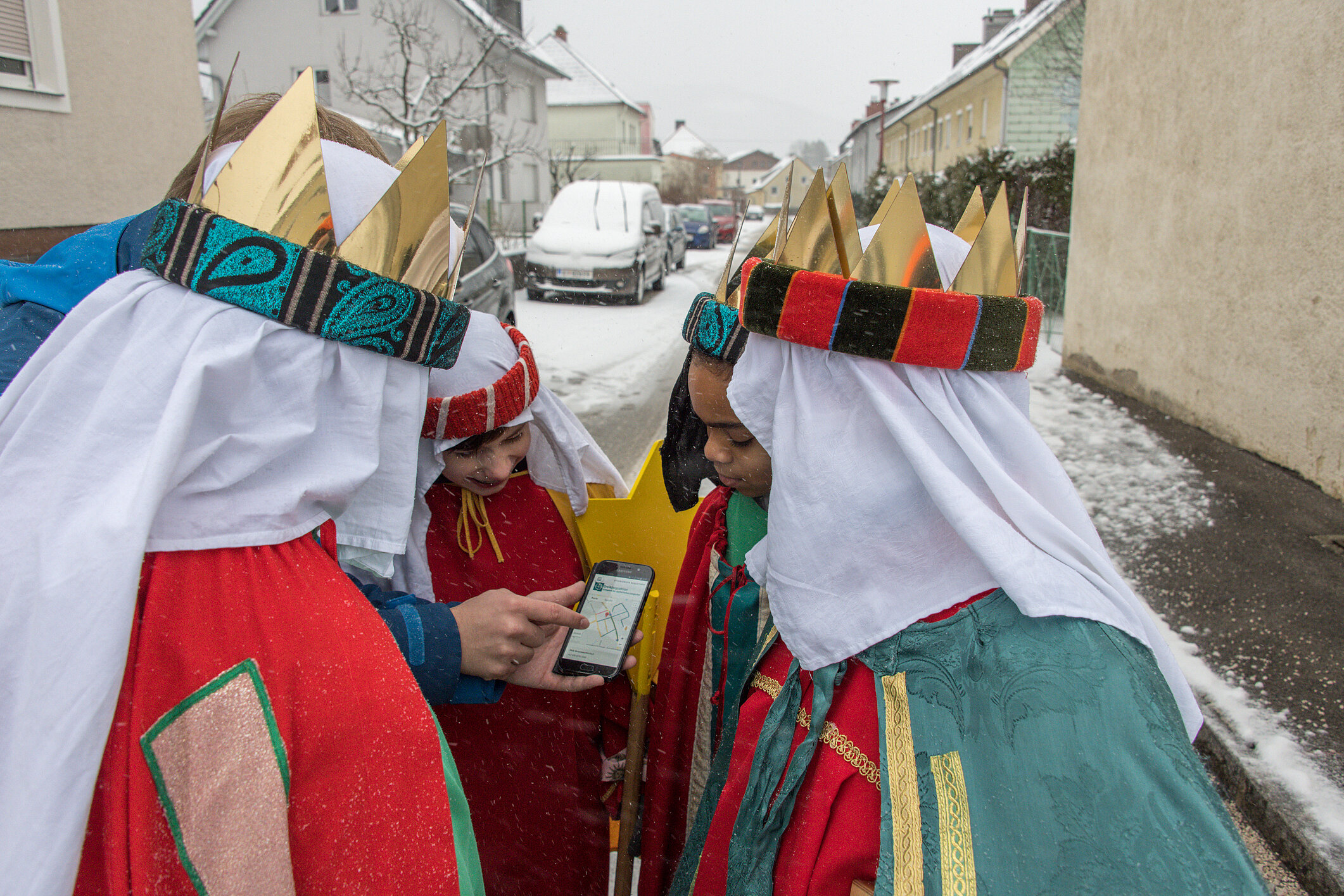 Vier als Sternsinger verkleidete Kinder stehen im Schnee und schauen gemeinsam auf ein Smartphone.