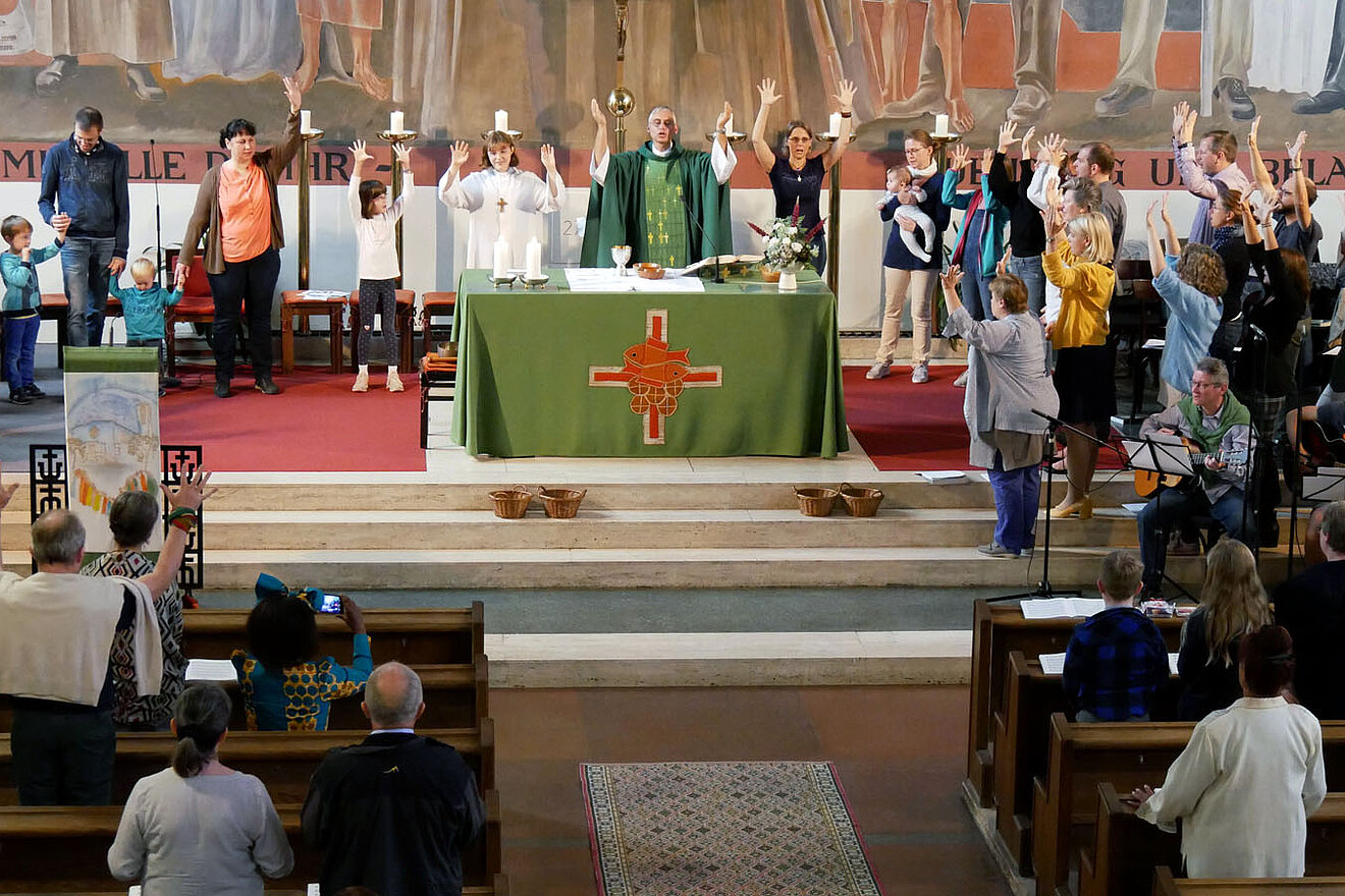 Spenden aus der thematischen Messe in der Pfarre Sandleiten kommen einem Projekt für Nahrungssicherheit in Tansania zu Gute. Foto: Lackermayer Der Altarraum der Pfarre Sandleiten (Wien) während einer Messfeier mit einigen Menschen, die die Arme hochhalten.
