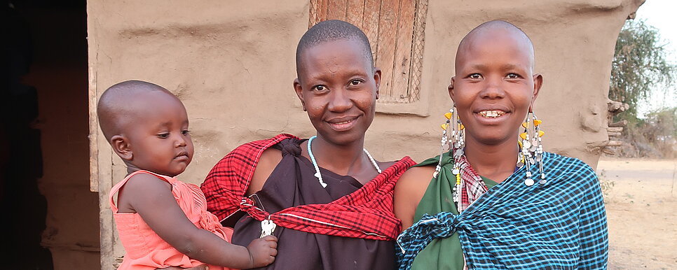 Zwei Frauen in traditioneller Kleidung stehen vor einem Lehmhaus. Die Frau links hält ein kleines Kind auf dem Arm, das in einem rosa Kleidchen gekleidet ist. Alle drei schauen lächelnd in die Kamera.