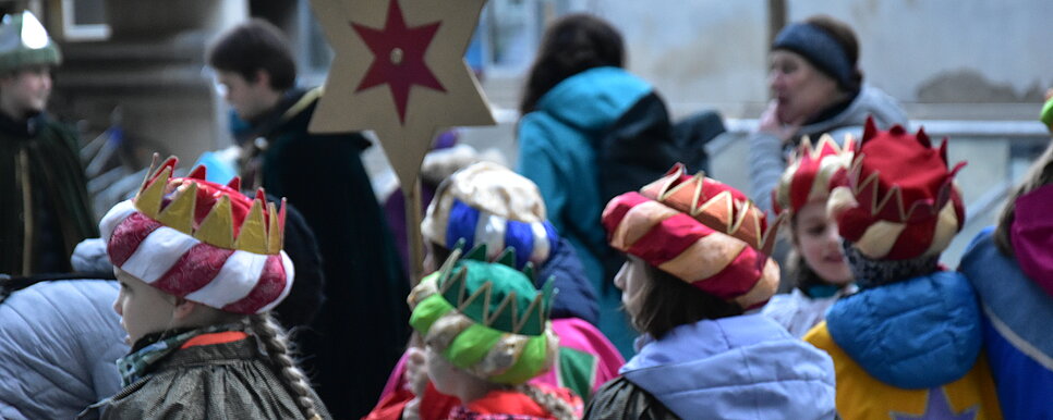 Das Bild zeigt eine Gruppe von Kindern, die als Sternsinger verkleidet sind. Sie tragen bunte Umhänge und Kronen sowie einen Stern, der auf einem Stab gehalten wird.