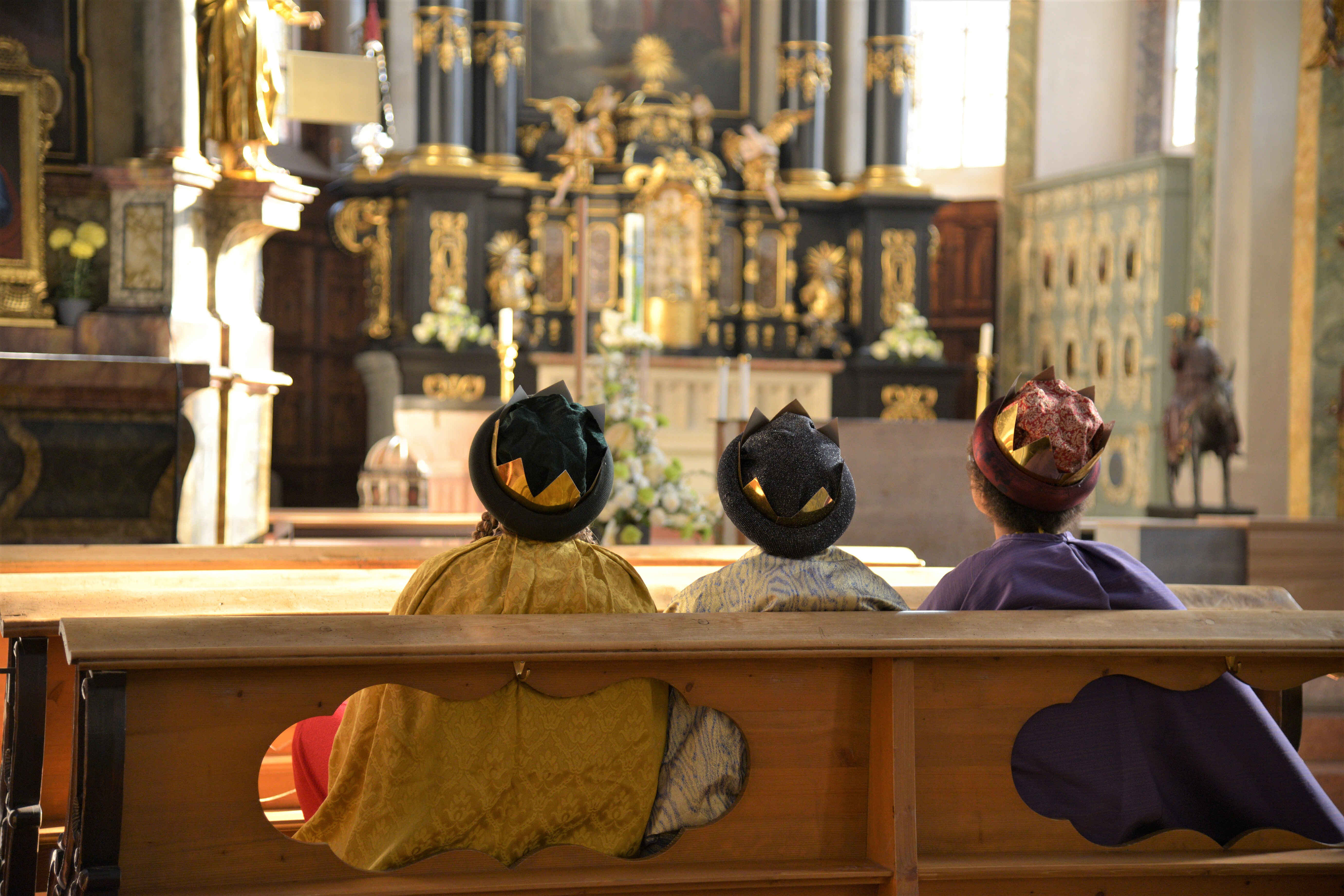 Drei Kinder sitzen im Königsgewand in einer Kirchenbank und schauen nach vorne.