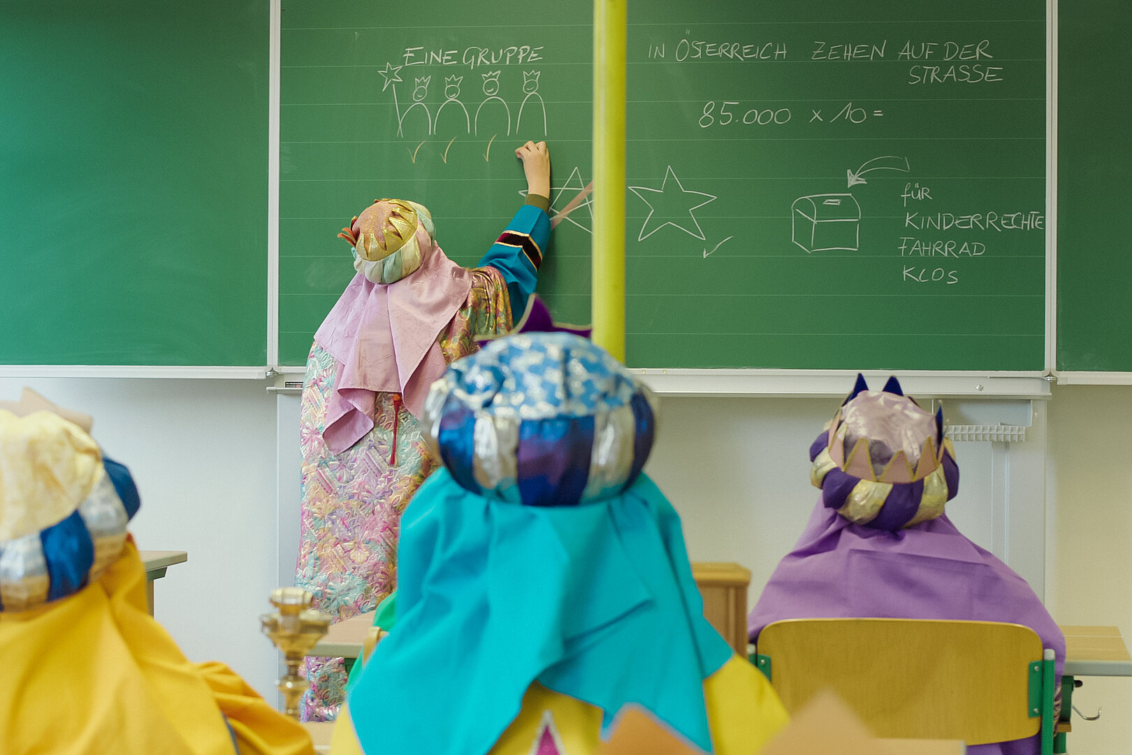 Ein Kind malt und schreibt etwas auf die Tafel, andere Kinder hören gespannt zu. Alle Kinder tragen Kleidung und Kronen der heiligen drei Könige.