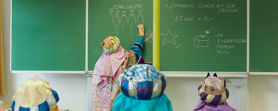 Ein Kind schreibt Informationen zum Sternsingen auf eine grüne Tafel und drei andere Kinder sitzen auf Stühlen und schauen zu.