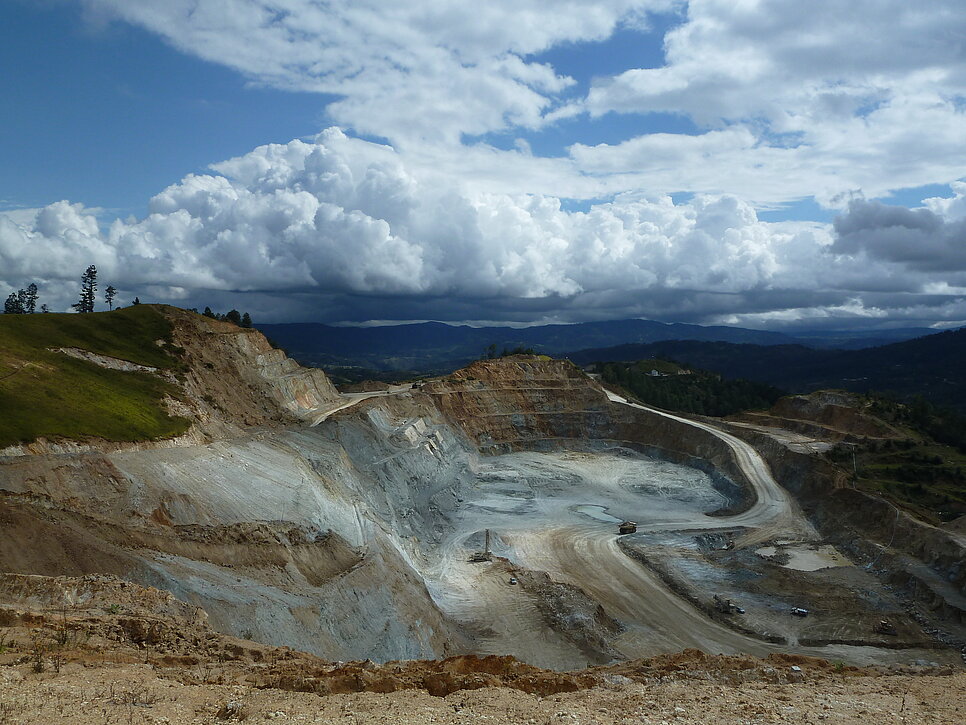 Das Bild zeigt eine große Goldmine im Tagebau in Guatemala, eingebettet in eine hügelige Landschaft. Der Boden ist tief ausgehoben, das Gestein wirkt karg und grau, einzelne Baufahrzeuge sind erkennbar. Über der Szene ziehen dichte Wolken am Himmel auf.