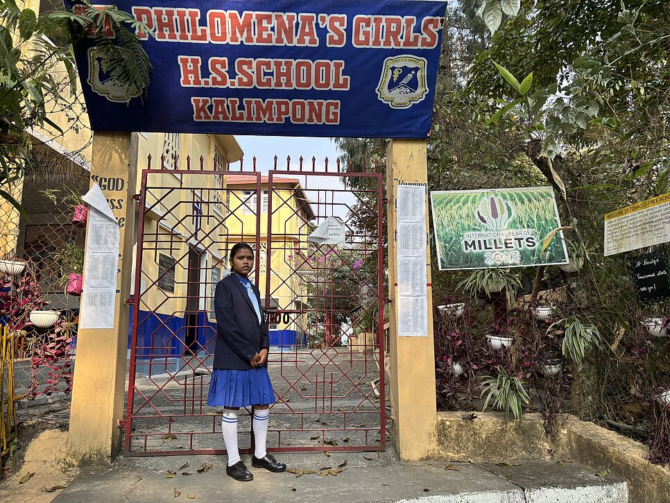 Ein Mädchen in Schuluniform mit blauem Rock und weißer Bluse steht vor einem roten Tor. Über dem Tor hängt ein blaues Schild mit der Aufschrift „Philomena’s Girls H.S. School Kalimpong“. Im Hintergrund sind gelbe Schulgebäude und Bäume zu sehen.