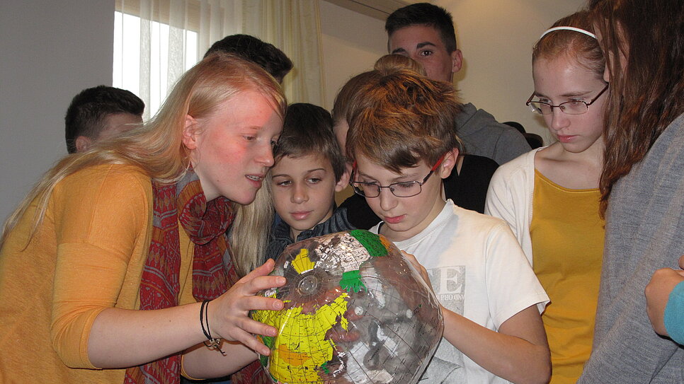 Mehrere Kinder und Jugendliche betrachten gemeinsam einen Globus. Sie zeigen interessiert auf verschiedene Länder und stehen dicht beieinander in einem hellen Raum.