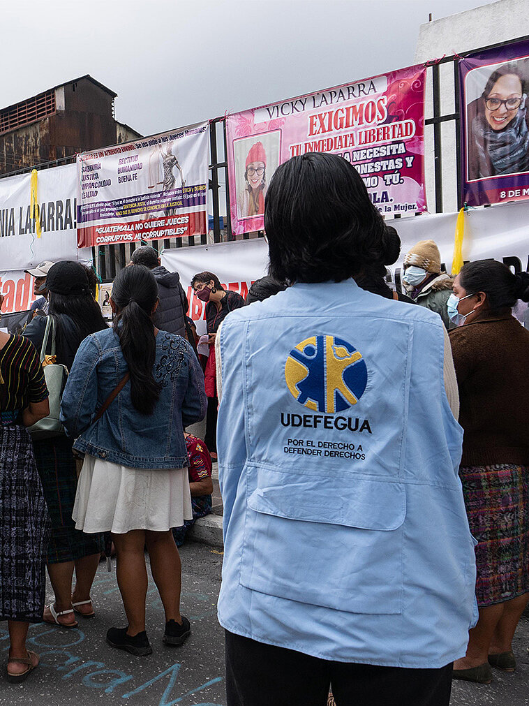 Im Bild sieht man eine Person mit einer hellblauen Jacke, auf deren Rücken das Logo von UDDFEGUA (Unidad de Defensores y Defensoras de Derechos Humanos en Guatemala) zu sehen ist. Diese Person steht inmitten einer Gruppe von Menschen, die sich offenbar auf eine Demonstration oder Versammlung konzentrieren. Im Hintergrund sind Banner und Plakate sichtbar, die für Gerechtigkeit und Menschenrechte werben.